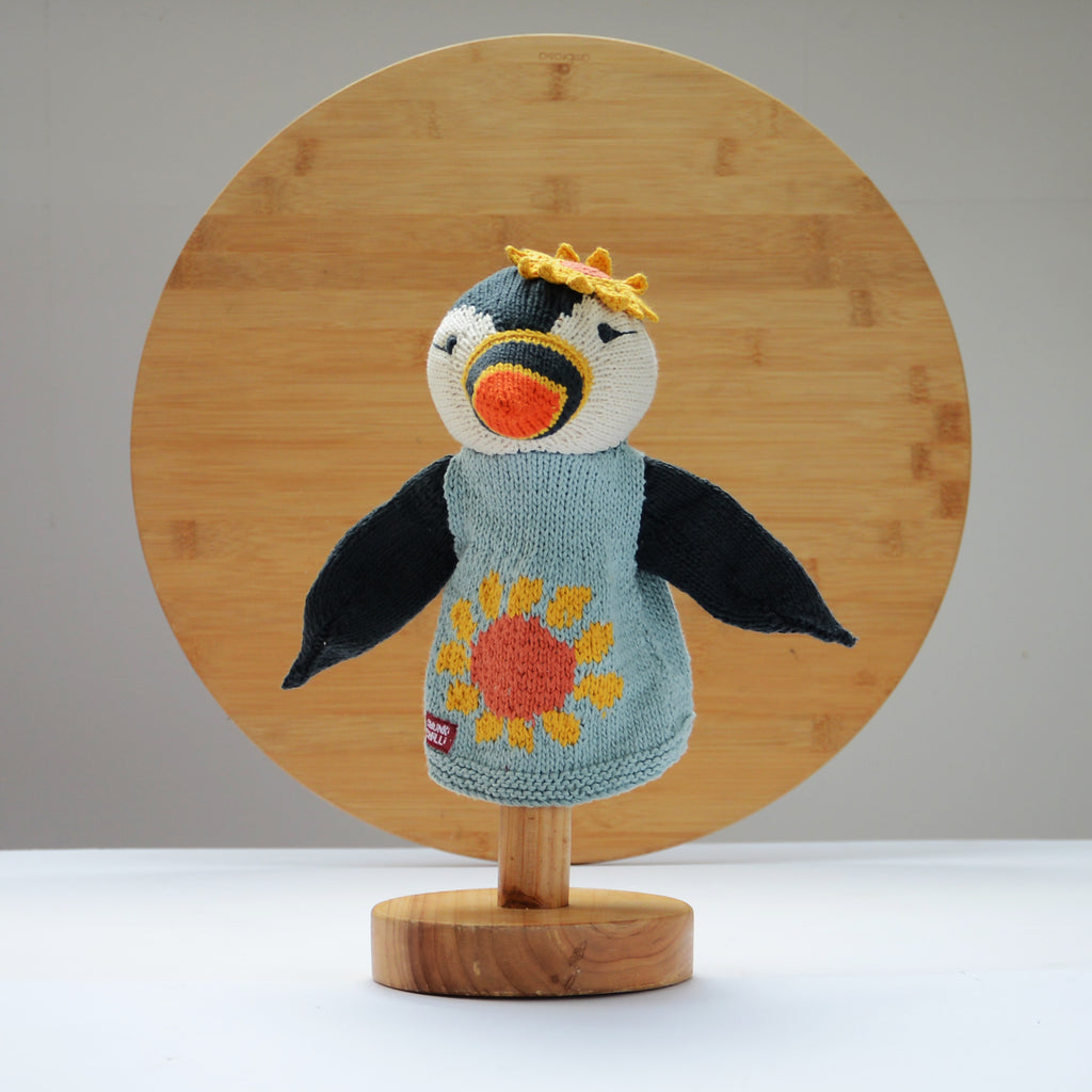 A charming knitted hand puppet by ChunkiChilli featuring a puffin with black wings, an orange beak, and a sunflower-patterned blue jumper, displayed on a wooden stand with a circular wooden background.